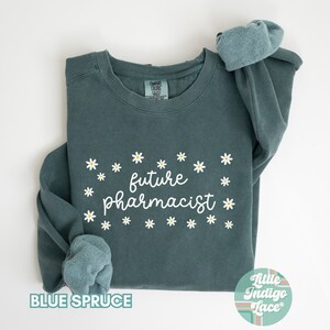 May include: A teal blue sweatshirt with the text "future pharmacist" in white cursive lettering surrounded by white daisy flowers. The sweatshirt is folded in half and the sleeves are rolled up. The text "BLUE SPRUCE" is printed in white on the bottom left corner of the image. The text "Little Indigo Lace" is printed in a colorful retro font on the bottom right corner of the image.