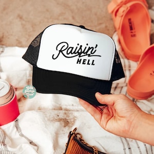 May include: A black and white trucker cap featuring a white front panel with the black text Raisin' HELL in a mix of script and block lettering. The cap has a black peak and black mesh side and back panels for ventilation and a structured fit.