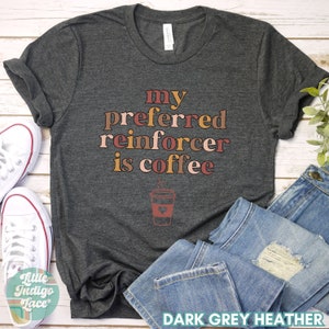May include: Dark grey heather t-shirt with the text "my preferred reinforcer is coffee" in brown, pink, and orange lettering. A stylized coffee cup with a heart and steam is below the text.