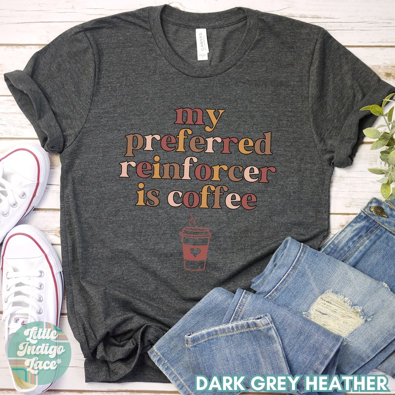 May include: Dark grey heather t-shirt with the text "my preferred reinforcer is coffee" in brown, pink, and orange lettering. A stylized coffee cup with a heart and steam is below the text.