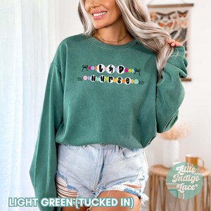 K&ouml;nnte beinhalten: Hellgr&uuml;ner Pullover mit dem Text "L&D Nurse" in schwarzen Buchstaben, umgeben von bunten Kreisen. Der Pullover ist in eine hellblaue Jeans-Shorts gesteckt.