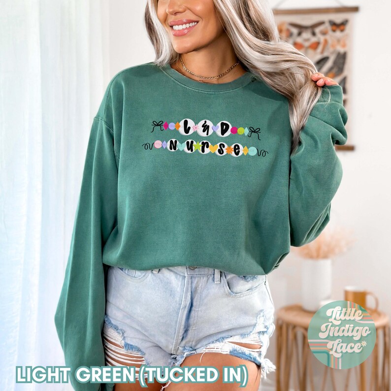 K&ouml;nnte beinhalten: Hellgr&uuml;ner Pullover mit dem Text "L&D Nurse" in schwarzen Buchstaben, umgeben von bunten Kreisen. Der Pullover ist in eine hellblaue Jeans-Shorts gesteckt.