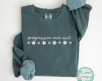 Op de afbeelding: Een tealblauwe trui met de tekst "progressive care unit" en een rij witte madeliefjes met gele centers. De trui is dubbelgevouwen en de mouwen zijn opgerold.