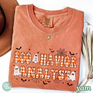 May include: A burnt orange short-sleeve t-shirt with a crew neckline. The front features the text "BOO HAVIOR ANALYST" in a wavy, orange and white checkered font, accompanied by illustrations of three small white ghosts, bats, and spiderwebs.