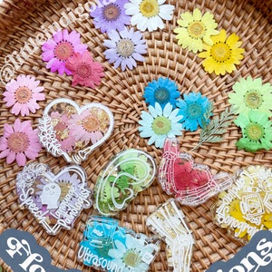 Pu&ograve; includere: Un cesto di vimini pieno di fiori pressati in vari colori, tra cui rosa, viola, blu, giallo e verde. I fiori sono disposti in un cerchio attorno al bordo del cesto. Al centro del cesto ci sono diversi pezzi di resina con diversi disegni, tra cui un cuore, un cervello e una mano. I pezzi di resina sono decorati con fiori pressati e scaglie dorate. Il testo "floral color options" &egrave; scritto su un adesivo grigio nell'angolo in basso a destra dell'immagine.