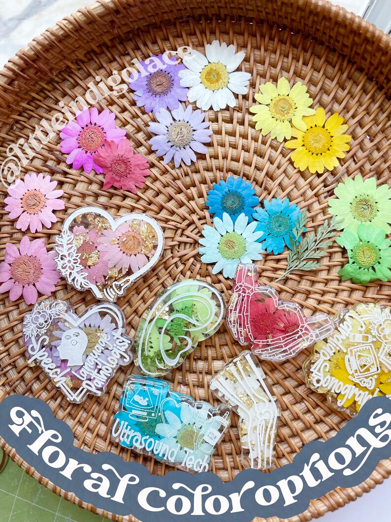 Pu&ograve; includere: Un cesto di vimini pieno di fiori pressati in vari colori, tra cui rosa, viola, blu, giallo e verde. I fiori sono disposti in un cerchio attorno al bordo del cesto. Al centro del cesto ci sono diversi pezzi di resina con diversi disegni, tra cui un cuore, un cervello e una mano. I pezzi di resina sono decorati con fiori pressati e scaglie dorate. Il testo "floral color options" &egrave; scritto su un adesivo grigio nell'angolo in basso a destra dell'immagine.