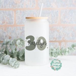 May include: A frosted glass water bottle with a bamboo lid and a green and black design that says "30 Katelyn" with a small flower and stars.