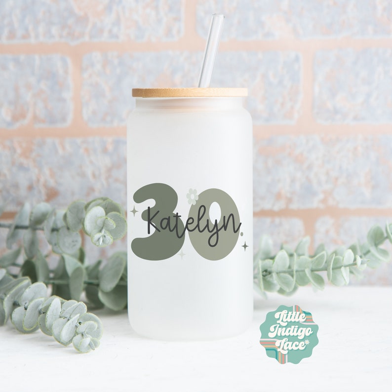 May include: A frosted glass water bottle with a bamboo lid and a green and black design that says "30 Katelyn" with a small flower and stars.