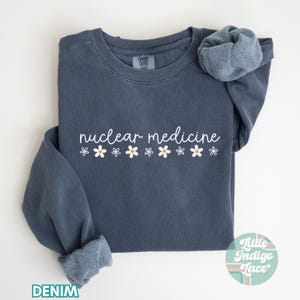 May include: A dark blue sweatshirt with the text "nuclear medicine" in white lettering and a row of white daisies with yellow centers.