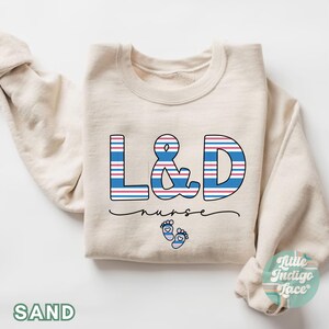 May include: A beige sweatshirt with the text "L&D nurse" in blue and red striped letters. The letters are outlined in black. There are two small blue footprints with red hearts below the text.