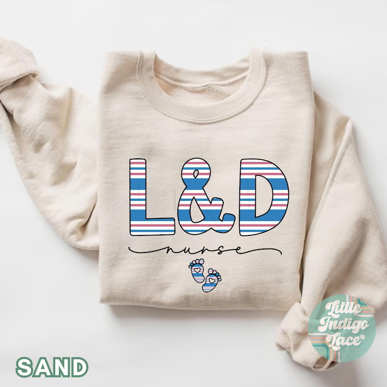 May include: A beige sweatshirt with the text "L&D nurse" in blue and red striped letters. The letters are outlined in black. There are two small blue footprints with red hearts below the text.