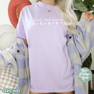 Puede incluir: Una camiseta de color morado claro con el texto "critical care nurse" en escritura blanca con una fila de peque&ntilde;as flores blancas debajo.