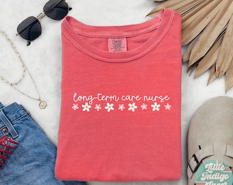 May include: A watermelon-colored Comfort Colors t-shirt with the words "long-term care nurse" in white script, accented by small daisy graphics. The shirt is displayed with sunglasses, jewelry, and denim shorts.