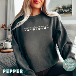 K&ouml;nnte beinhalten: Ein dunkelgrauer Pullover mit dem Text "pediatric intensive care unit" und einer Reihe wei&szlig;er G&auml;nsebl&uuml;mchen, die auf der Vorderseite aufgedruckt sind. Der Pullover wird von einer Person mit langen blonden Haaren getragen.