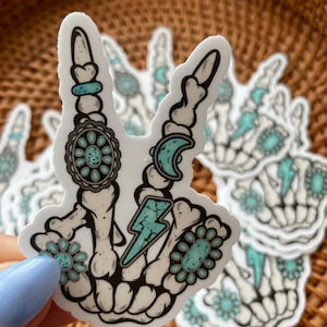 May include: A white sticker featuring a skeleton hand in a peace sign gesture. The design includes turquoise accents, a crescent moon, and a lightning bolt. The sticker is decorated with floral and circular patterns.