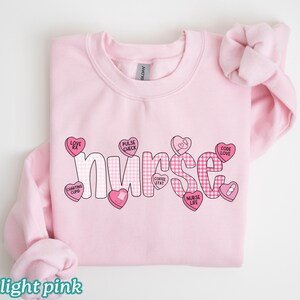 May include: Light pink sweatshirt with the word "nurse" in a playful font, decorated with pink hearts and candy hearts. The candy hearts have phrases like "Love Rx" and "Code Love". The sweatshirt is made of a soft material.