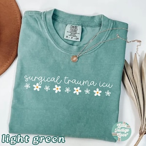 May include: Light green t-shirt with the words "surgical trauma icu" in white script, accented with white daisy graphics. A silver necklace with a pendant rests on the shirt. The shirt is labeled "Comfort Colors".