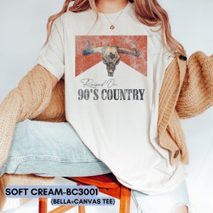 May include: A white t-shirt with a distressed red and white graphic of a bull skull with the text "Raised On 90's Country".