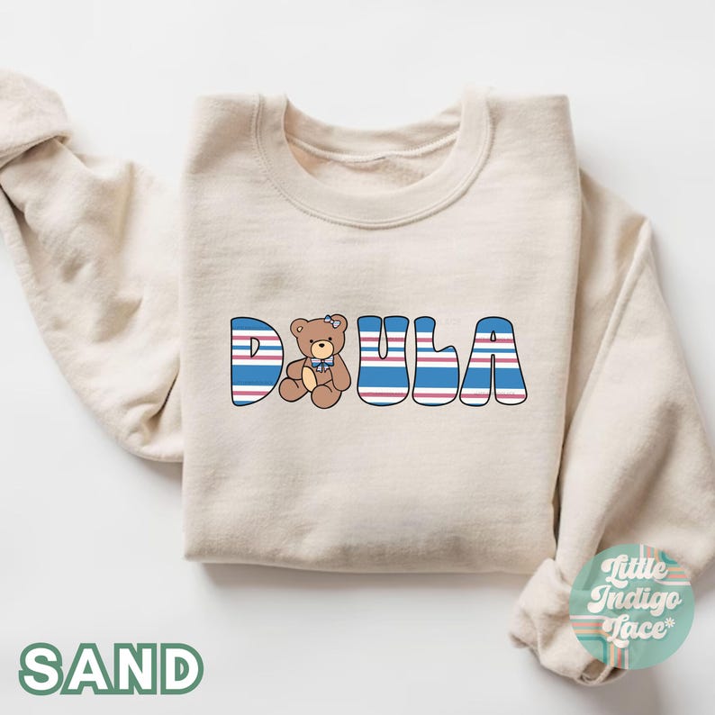 May include: A cream-colored sweatshirt with the word "DOULA" spelled out in blue and red striped letters. A small brown teddy bear with a blue bow is sitting in the middle of the word.
