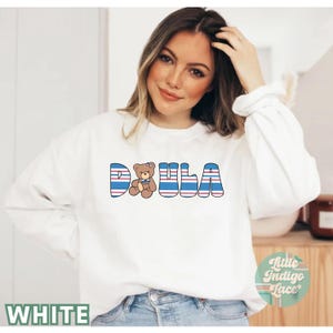 May include: White sweatshirt with blue and red stripes spelling out "DOULA" with a brown teddy bear wearing a blue bow in the middle of the word.
