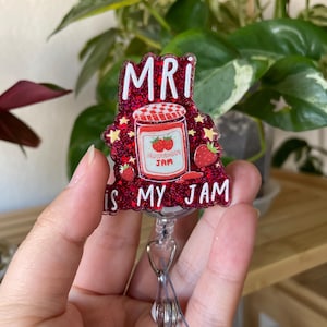 May include: A red glitter badge reel with the text "MRI IS MY JAM". The badge features a strawberry jam jar illustration. The reel has a clear plastic strap and a metal clip.