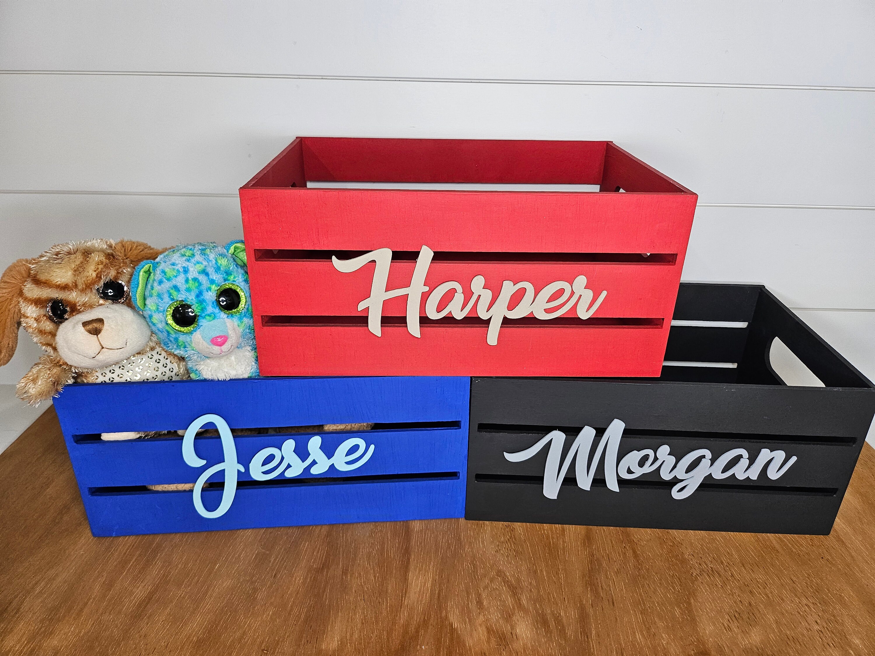 Three personalized wooden boxes. Names are hand-painted in different colors: beige, light blue, and gray. The boxes are red, black and royal blue. Ideal for a personalized gift or adding a decorative touch to a nursery or kids room.