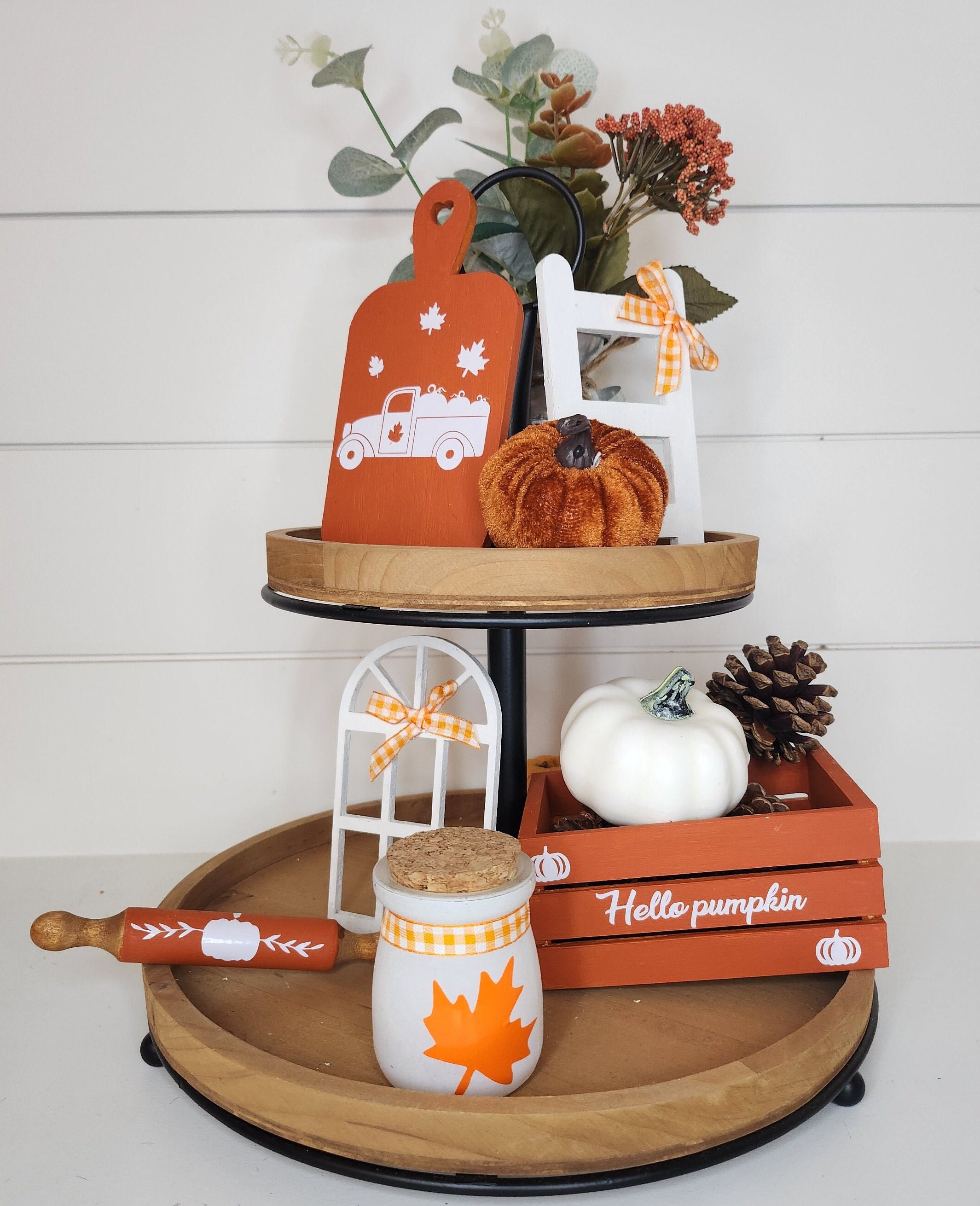 Two-tiered wooden stand with fall decor including an orange mini crate with custom vinyl inscription, a jar with a maple leaf, an orange cutting board with white custom vinyl design, orange rolling pin, a white mini window and small rustic ladder.