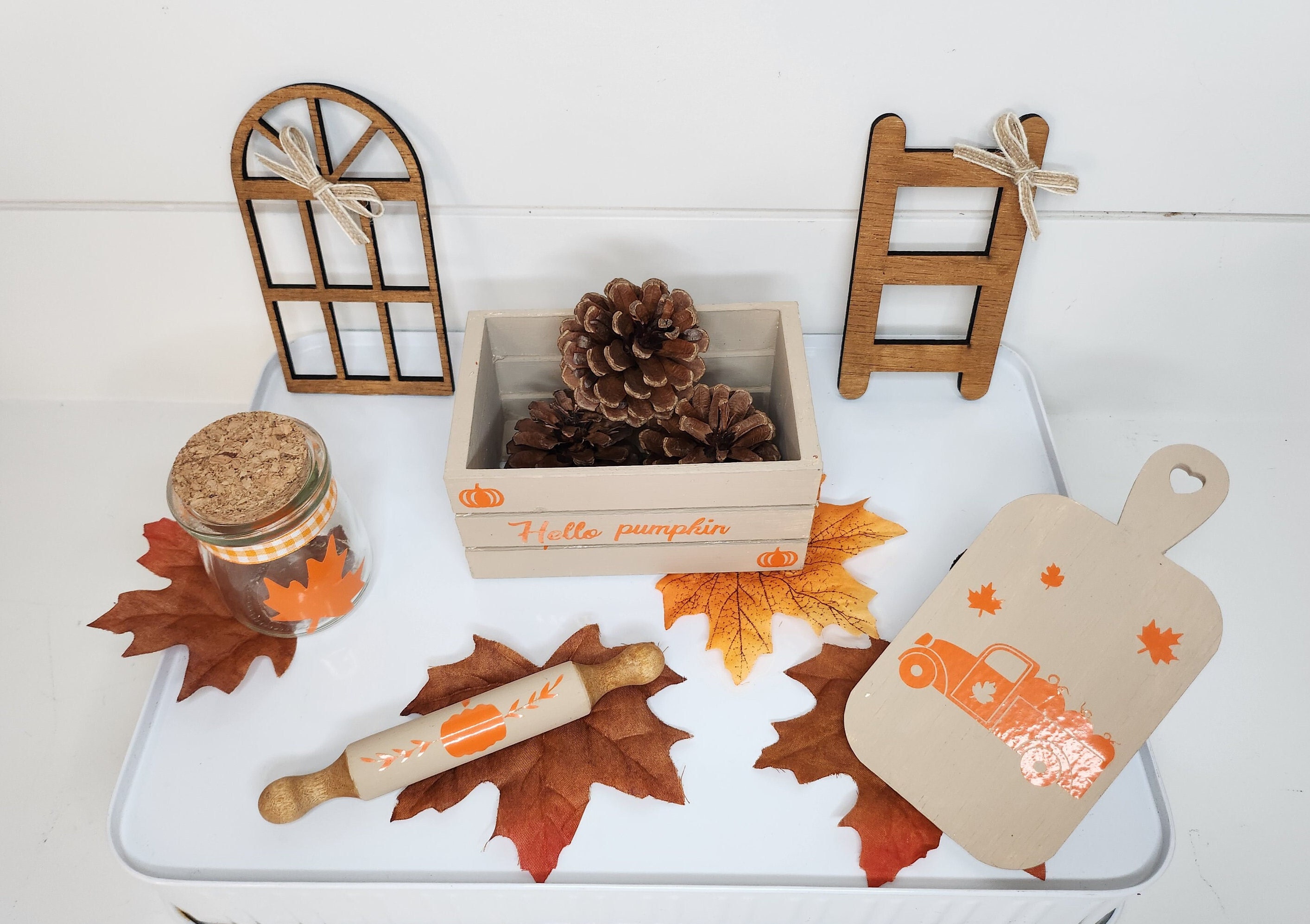 White tray with fall decor including a beige mini crate with custom vinyl inscription, a jar with a maple leaf, a beige cutting board with orange custom vinyl design, beige rolling pin, stained mini window and small rustic ladder.