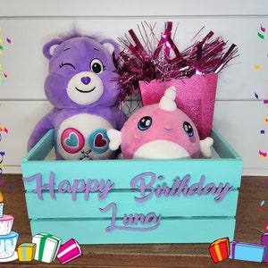 May include: A turquoise wooden crate with the words "Happy Birthday Luna" painted on the front. The crate is filled with a purple Care Bear plush toy, a pink narwhal plush toy, and a pink and silver streamer. There are also colorful confetti and wrapped gifts around the crate.