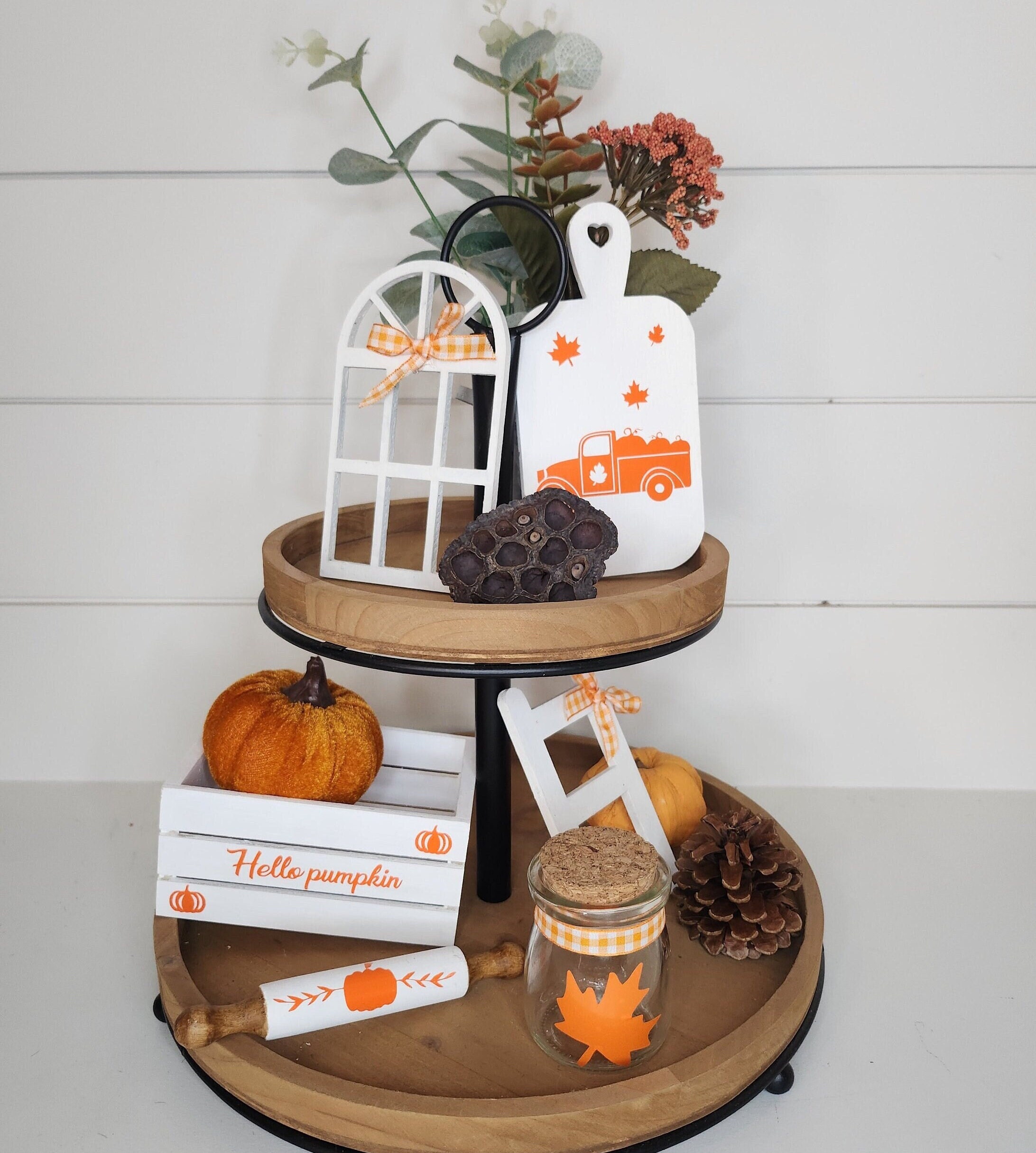 Two-tiered wooden stand with fall decor including a white mini crate with custom vinyl inscription, a jar with a maple leaf, a white cutting board with orange custom vinyl design, white rolling pin, a white mini window and small rustic ladder.