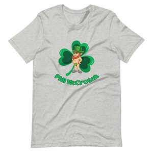 Op de afbeelding: Een grijs T-shirt met een groene klaver en een cartoon-kabouter met een groene hoed. De tekst "Phil McCrotch" is onder de klaver gedrukt.