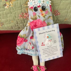 May include: A fabric ghost doll with a light blue floral pattern, white daisy trim, and pink shoes, holding a card labelled Ghostie Pocket Hug with a small white ghost charm featuring a mint green hat and orange accent.