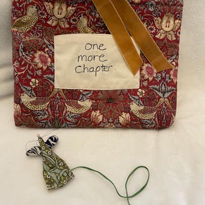 May include: A red patterned book cover with a floral and bird design, a cream patch with the text "one more chapter", and a gold ribbon. A small fabric angel bookmark is below.