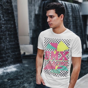 May include: A white t-shirt with a vintage-style graphic design featuring a checkered background, colorful geometric shapes, and the text "BMX RACING 1969".