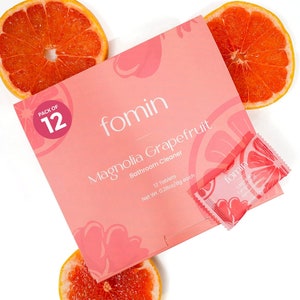 May include: A pink box of "fomin Magnolia Grapefruit Bathroom Cleaner" with a "pack of 12" label. The box is surrounded by grapefruit slices. A single packet of the cleaner is visible.