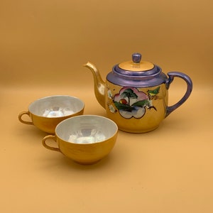 May include: A gold and purple teapot with floral designs and two matching teacups. The teapot has a lid and a handle. The teacups have a white interior.