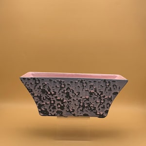 May include: A rectangular planter with a textured, speckled exterior in shades of gray and black. The interior rim is a soft pink. The planter is displayed on a clear stand, showcasing its unique design.