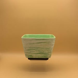 May include: A square, light green planter with a textured, horizontal striped pattern. The interior is a solid light green. The planter sits on a black base, and is displayed against a light brown background.
