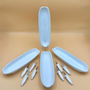May include: A set of three white ceramic corn-shaped serving dishes with textured kernels and husks, accompanied by six small metal corn picks featuring white ceramic handles shaped like miniature corn cobs.