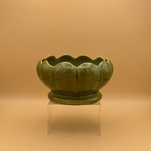 May include: A green ceramic planter with a scalloped rim. The planter sits on a clear acrylic stand against a tan background. The planter has a round base and a textured surface.