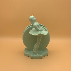May include: A light green ceramic vase shaped like a ballerina. The ballerina is standing on a circular base and is holding her arms out to the side. The vase is a vintage style.