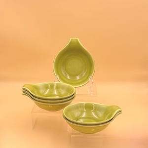 May include: A set of five small, round, light green ceramic bowls with a single handle on one side. The bowls feature a smooth, glossy finish and a uniform color throughout. They are arranged in two stacks, with one bowl standing upright behind.