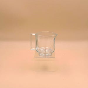 May include: A clear glass creamer pitcher featuring a cylindrical body, a small pouring spout, and a square-shaped handle. The pitcher is transparent, showing a smooth surface and a thick base, designed for serving liquids like cream or milk.
