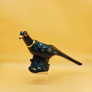 May include: A glossy black ceramic bird figurine featuring gold-painted accents on the beak, neck, wings, and tail. The bird is perched on a textured black base with gold detailing, showcasing a sleek, elongated form and a long, pointed tail.