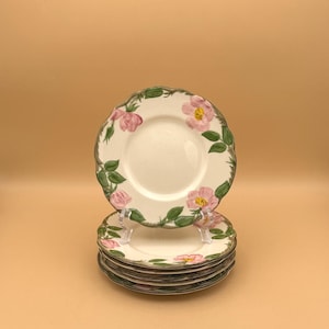 May include: A stack of white ceramic plates with a floral design. Each plate features pink flowers with yellow centers and green leaves around the rim. The plates are stacked on a clear plastic stand against a light orange background.