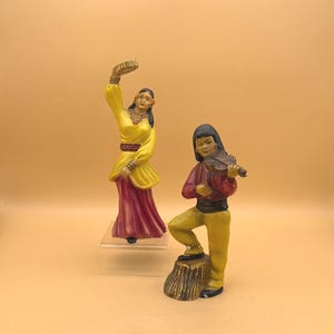 May include: Two ceramic figurines depicting traditional dancers. One figure is in a yellow dress with a red skirt, arms raised. The other figure is playing a violin, standing on a tree stump, wearing a red shirt and yellow pants.