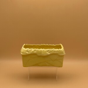 May include: A yellow ceramic planter with a textured, folded design. The planter is sitting on a clear acrylic stand.
