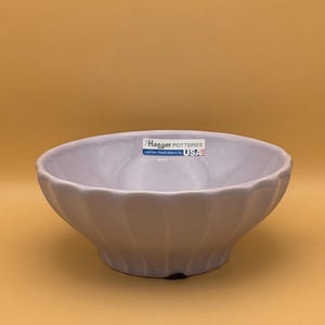 May include: A light purple ceramic bowl with a fluted design. The bowl is made by Haeger Potteries and is marked "Lead Free - Proudly Made in the USA".