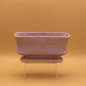 May include: A lavender ceramic planter with a rectangular shape and a matching rectangular base. The planter features a wavy, textured design on the sides and a row of small dots along the top and bottom edges.