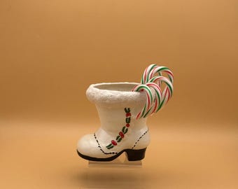 May include: A white ceramic boot-shaped container with a decorative floral design in red and green. The boot is filled with red, white, and green candy canes. The boot has a black heel and is on a clear stand.